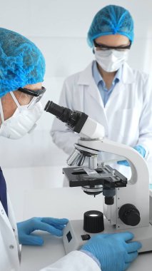 Mavi koruyucu saç filesi, maske ve gözlüklü iki bilim adamı bir laboratuarda mikroskop altında araştırma yapıyorlar. Tıp ve bilim kavramı.