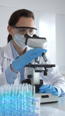 Koruyucu gözlük takan ve maske takan kadın bilim adamı mikroskop kullanarak laboratuvarda numuneleri analiz ediyor mavi sıvı test tüpleriyle çevrili yeni ilaç veya aşıları araştırıyor.