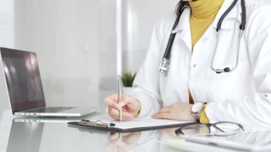 Beyaz önlüklü ve steteskoplu bir doktor bir not panosuna not yazıyor ve hasta bilgilerini kontrol ediyor. Tıp, sağlık ve bilim konsepti