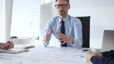 Gözlüklü güvenilir işadamı bir şirket toplantısına liderlik ediyor, mali tabloları gözden geçiriyor, veri içgörülerini açıklıyor ve iş arkadaşlarıyla stratejik büyüme fırsatlarını tartışıyor..