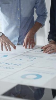 İş arkadaşları mali raporları inceliyor ve masadaki çizelge ve grafiklere işaret ediyor. Verileri analiz etmek ve stratejileri planlamak için birlikte çalışıyorlar.