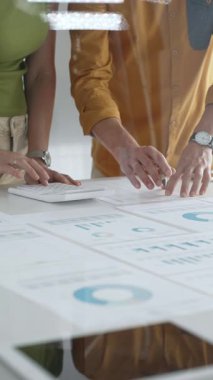 İki iş adamı bir proje üzerinde işbirliği yapıyor, bir tabloda gösterilen finansal çizelgeleri ve istatistikleri analiz ediyorlar, kesin hesaplamalar için bir hesap makinesi kullanıyorlar