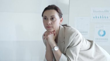 İş kadını ellerini sıkarak masaya yaslanıyor ve arka planda grafikler ve çizelgeler olan parlak, modern bir ofiste bir sonraki iş hamlesini düşünüyor. İş adamları konsepti