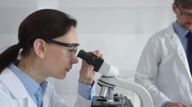 Laboratuvarda araştırma sırasında mikroskoptan bakan koruyucu gözlük takan kadın bir bilim adamı ve arka planda çalışan erkek arkadaşı. Tıp ve bilim konsepti.