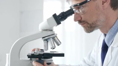 Yetişkin erkek bilim araştırmacısı mikroskopla örnek analiz ediyor, yüksek teknoloji laboratuvar ortamında koruyucu giysiler giyiyor. Sağlık, tıp ve bilim konsepti.