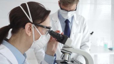 Laboratuvar önlüklü ve güvenlik teçhizatlı iki bilimadamı araştırma yapıyor. Biri mikroskopla gözlem yapıyor diğeri de elinde turkuaz sıvıyla test tüpü tutuyor. Tıp ve bilim konsepti.