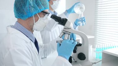 Koruyucu maske takan ve gözlük takan erkek bilim adamı mikroskop kullanarak laboratuvarda araştırma yapıyor ve elinde mataralar olan bir bilim adamı asistanı var. Sağlık ve bilim konsepti.