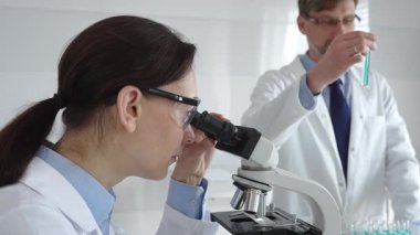 Laboratuvarda araştırma sırasında mikroskoptan bakan koruyucu gözlük takan kadın bir bilim adamı ve arka planda çalışan erkek arkadaşı. Tıp ve bilim konsepti.