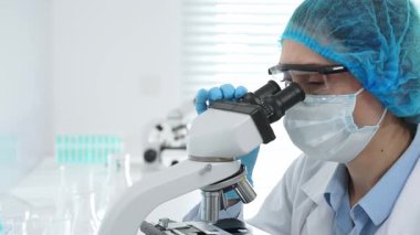 Kadın bilim adamı mikroskop ile numune analiz ediyor, tıbbi araştırma sırasında koruyucu laboratuvar giysileri giyiyor. Tıp, laboratuvar ve bilim konsepti.