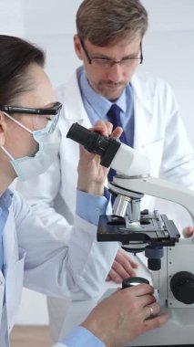 Koruyucu gözlük ve maske takan kadın bilim adamı, laboratuvarda tıbbi araştırma sırasında mikroskoptan bakıyor ve erkek meslektaşı arka planda çalışıyor. Tıp ve Bilim