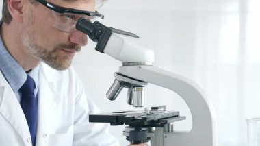 Yetişkin erkek bilim araştırmacısı mikroskopla örnek analiz ediyor, yüksek teknoloji laboratuvar ortamında koruyucu giysiler giyiyor. Sağlık, tıp ve bilim konsepti.