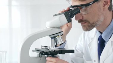 Yetişkin erkek bilim araştırmacısı mikroskopla örnek analiz ediyor, yüksek teknoloji laboratuvar ortamında koruyucu giysiler giyiyor. Sağlık, tıp ve bilim konsepti.
