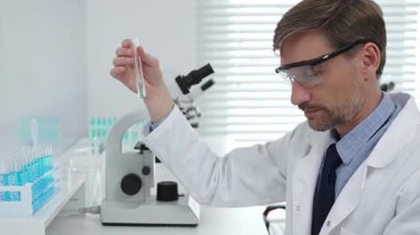Beyaz önlüklü ve koruyucu gözlüklü bir erkek bilim adamı laboratuvar ortamında test tüpündeki sıvıyı analiz ediyor. Tıp, sağlık ve bilim konsepti.