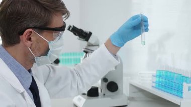 Koruyucu giysi giyen bilim araştırmacısı modern laboratuvar çalışma alanında mavi sıvı numunesini test tüpünden inceliyor. Tıp, sağlık ve bilim konsepti