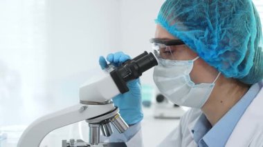 Kadın bilim adamı mikroskop ile numune analiz ediyor, tıbbi araştırma sırasında koruyucu laboratuvar giysileri giyiyor. Tıp, laboratuvar ve bilim konsepti.