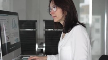 İş kadını verileri yönetir ve çağdaş bir ofiste bilgisayar ekranında finansal grafikleri analiz eder, raporlara ve iş stratejisine odaklanır, üretkenliği ve kurumsal başarıyı temsil eder