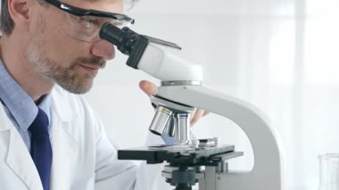 Yetişkin erkek bilim araştırmacısı mikroskopla örnek analiz ediyor, yüksek teknoloji laboratuvar ortamında koruyucu giysiler giyiyor. Sağlık, tıp ve bilim konsepti.