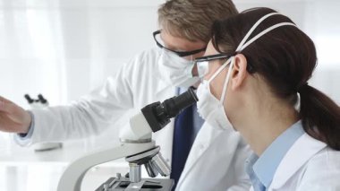 Laboratuvar önlüklü ve güvenlik teçhizatlı iki bilimadamı araştırma yapıyor. Biri mikroskopla gözlem yapıyor diğeri de elinde turkuaz sıvıyla test tüpü tutuyor. Tıp ve bilim konsepti.