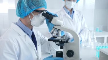 Koruyucu maske takan ve gözlük takan erkek bilim adamı mikroskop kullanarak laboratuvarda araştırma yapıyor ve elinde mataralar olan bir bilim adamı asistanı var. Sağlık ve bilim konsepti.