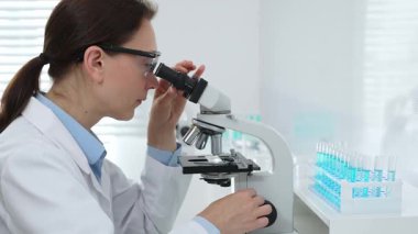Laboratuvar önlüğü ve koruyucu gözlüklü kadın bilim adamı modern bir laboratuvarda mikroskop altında test örnekleri inceliyor. Tıp, sağlık ve bilim konsepti