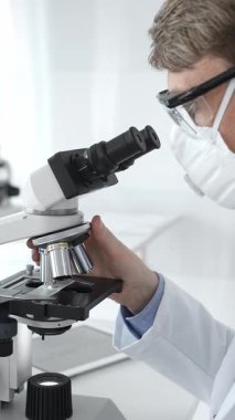 A researcher wearing a protective mask examines a sample under a microscope, conducting a detailed laboratory study. Concept of medicine and science.
