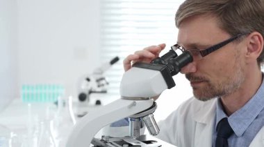 Beyaz önlük ve gözlük giyen erkek bilim adamı mikroskop kullanıyor ve modern bir laboratuvarda bilimsel araştırma yapıyor. Tıp, sağlık ve bilim konsepti.