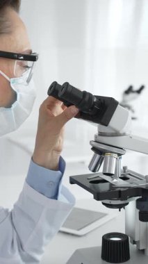 Koruyucu maske takan ve gözlük takan kadın bilim adamı mikroskopla numuneleri dikkatle inceliyor ve modern laboratuvarda araştırma yapıyor. Tıp ve bilim konsepti.