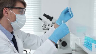 Laboratuvar önlüklü, koruyucu gözlüklü ve mavi eldivenli maskeli erkek bilim adamı modern laboratuardaki test tüplerine mikroskop ve test tüpleri rafı koyuyor. Tıp, sağlık ve bilim konsepti.