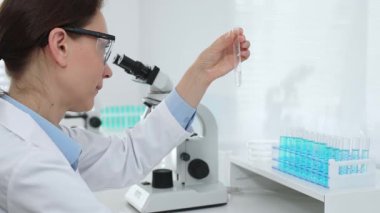 Laboratuvar önlüğü giyen ve koruyucu gözlük takan kadın bilim adamı modern laboratuarda mikroskop ve test tüpleri askısı ile test tüpünü temiz sıvıyla analiz ediyor. Tıp, sağlık ve bilim konsepti