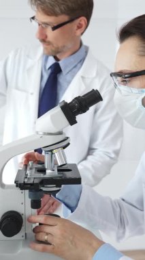 Koruyucu gözlük ve maske takan kadın bilim adamı, laboratuvarda tıbbi araştırma sırasında mikroskoptan bakıyor ve erkek meslektaşı arka planda çalışıyor. Tıp ve Bilim.