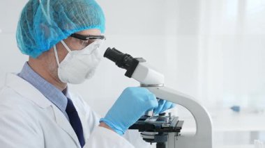 Üst düzey erkek bilim adamı koruyucu maske, saç filesi, gözlük ve eldiven takıyor mikroskop kullanarak tıbbi bir laboratuarda bilimsel araştırma yapıyor. Laboratuvar ve bilim konsepti