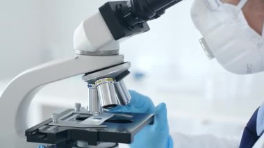 Mavi eldiven giyen bilim araştırmacısı mikroskop altında numuneleri dikkatlice analiz ederek hassas laboratuvar incelemesi yapıyor. Sağlık ve bilim konsepti.