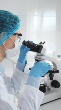 Mavi koruyucu giysiler giyen kadın bilim adamı mikroskop kullanarak laboratuvar ortamında örnekler inceleyerek bilimsel araştırma ve analiz yapıyor. Tıp ve bilim konsepti.