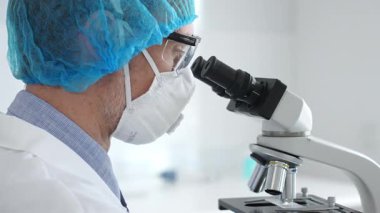 Koruyucu ekipman takan erkek bilim adamı mikroskop kaydırağını inceliyor, steril laboratuvar ortamında araştırma örneklerini analiz ediyor. Sağlık ve bilim konsepti.