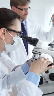 Koruyucu gözlük ve maske takan kadın bilim adamı, laboratuvarda tıbbi araştırma sırasında mikroskoptan bakıyor ve erkek meslektaşı arka planda çalışıyor. Tıp ve Bilim