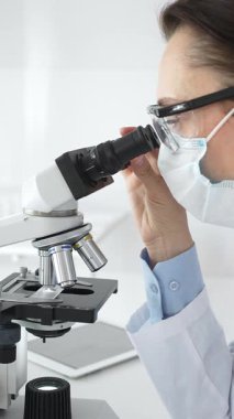 Koruyucu maske takan ve gözlük takan kadın bilim adamı mikroskopla numuneleri dikkatle inceliyor ve modern laboratuvarda araştırma yapıyor. Tıp ve bilim konsepti.