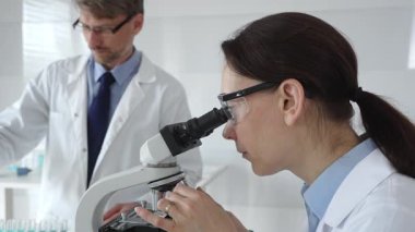 Laboratuvarda araştırma sırasında mikroskoptan bakan koruyucu gözlük takan kadın bir bilim adamı ve arka planda çalışan erkek arkadaşı. Tıp ve bilim konsepti.