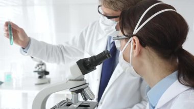 Laboratuvar önlüklü ve güvenlik teçhizatlı iki bilimadamı araştırma yapıyor. Biri mikroskopla gözlem yapıyor diğeri de elinde turkuaz sıvıyla test tüpü tutuyor. Tıp ve bilim konsepti.