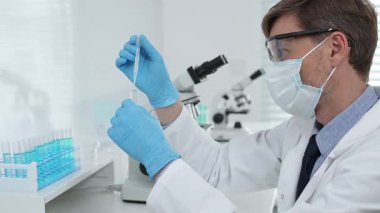 Laboratuvar önlüklü, koruyucu gözlüklü ve mavi eldivenli maskeli erkek bilim adamı modern laboratuardaki test tüplerine mikroskop ve test tüpleri rafı koyuyor. Tıp, sağlık ve bilim konsepti.