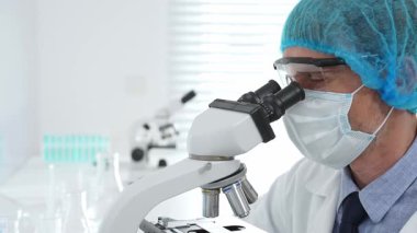 Kıdemli bilim adamı mikroskopla numune inceliyor, steril laboratuvar ortamında tıbbi araştırma yaparken koruyucu giysiler giyiyor. Tıp, sağlık ve bilim konsepti