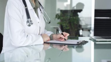 Masasında steteskop yazılı beyaz önlüklü doktor muayene sırasında hasta bilgilerini belgeliyor. İlaç ve sağlık hizmetleri kavramı