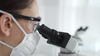 Koruyucu maske takan ve gözlük takan kadın bilim adamlarının mikroskop altında örnekleri dikkatle inceleyerek modern laboratuvarda araştırma yapmalarına dikkat edin. Tıp ve bilim konsepti.