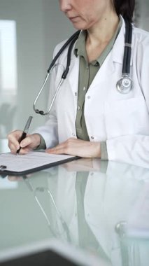 Masasında steteskop yazılı beyaz önlüklü doktor muayene sırasında hasta bilgilerini belgeliyor. İlaç ve sağlık hizmetleri kavramı