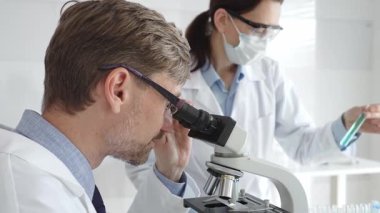 Bilimsel uzmanlar mikroskopla numuneleri inceliyor. Araştırma sırasında her ikisi de beyaz laboratuvar önlüğü giymiş koruyucu maske takan laboratuvar asistanı. Tıp ve Bilim.