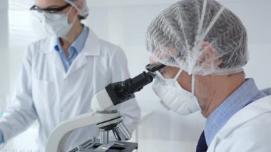 Koruyucu maske takan kıdemli erkek bilim adamı, gözlük ve saç filesi takıyor, mikroskop kullanarak genç bayan meslektaşlarına yardım ediyor, tablet tutuyor ve laboratuvarda notlar alıyor. Tıp ve bilim konsepti.