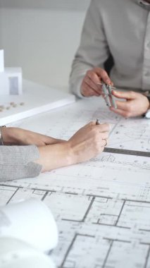 Mimarlar detaylı planlar ve beyaz bir masanın üzerinde bir ölçek modeli etrafında toplanarak, modern bir konut projesinde işbirliği yaparken planları analiz ediyor, araçlar ve belgeler çiziyor ve kapatıyorlar.