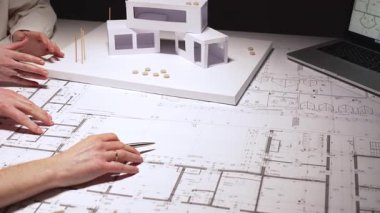 Mimarlar planlar ve ölçek modeli, cetvel ve kalemle planları ölçme ve işaretleme konusunda işbirliği yaparken, modern bir ofis iş yerindeki dizüstü bilgisayar ve evraklara atıfta bulunuyorlar.