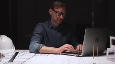 Stresli mimar baş ağrısı ve hayal kırıklığı hissediyor, gece geç saatlere kadar karanlık bir ofiste çalışıyor, planlar, proje teslim tarihleri ve fazla mesai talep ediyor.