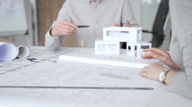 Bir konut projesinde işbirliği yapan mimarlar, planlar ve ölçekli bir modeli bir masada inceleyerek, tasarım detaylarını, inşaat planlarını ve gelecekteki gelişimi tartışarak,