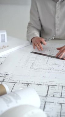 Mimarlar detaylı planlar ve beyaz bir masanın üzerinde bir ölçek modeli etrafında toplanarak, modern bir konut projesinde işbirliği yaparken planları analiz ediyor, araçlar ve belgeler çiziyor ve kapatıyorlar.